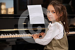 Cute child girl pianist sitting at grand piano, enjoys the rhythm of classical music while plays piano, smiles at camera