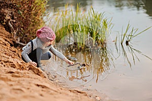 Cute child girl in pink knitted hat plays with stick on river side with sand beach