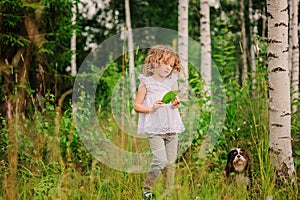 Cute child girl playing with leaves in summer forest with her dog. Nature exploration with kids.