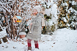 Cute child girl puts seeds in bird feeder in winter snowy garden