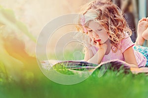 Cute child girl reading book and dreaming in summer sunny garden