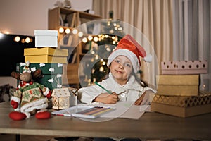 Cute child girl in red hat writes the letter to Santa Claus sitting at table near Christmas tree