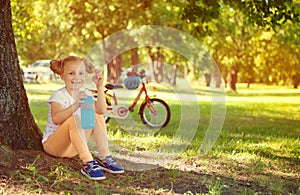 Cute child girl sit under the tree with bottle in hand. Bicycle