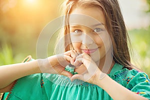 Cute child, little girl showing heart gesture and smiling outdoor. Love summer. Beautiful seven years old child enjoying nature