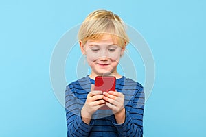 Cute child looking at his smartphone smiling. Boy playing video games on his phone, isolated over pastel blue background.