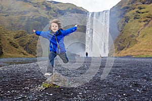 Cute child running in front of the Skogafoss waterfall in Iceland on a sunset cloudy day
