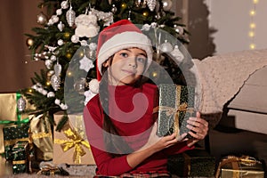 Cute child in Santa hat holding gift box near Christmas tree at home