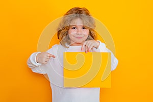 Cute child showing index finger on sheet of paper, isolated on yellow background. Portrait of a kid holding a blank