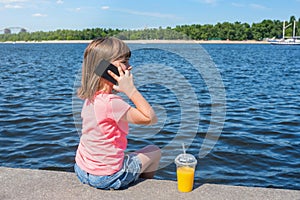 A cute child sits at the river and speaks by phone, in sunny day. View from a back