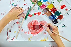 Cute child sitting at the table and drawing red heart on white paper