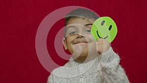 Cute child smiling and expressing his happinesswith green happy emoyi. Isolated on red background, slow motion