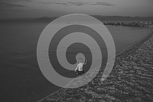 Cute child waving hand at seashore in evening. Boy sits in sea water, rear view. Farewell and parting concept. Toddler