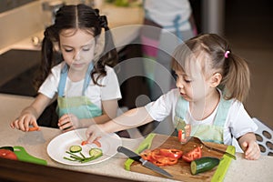 Cute children helping mother at kitchen. Adorable sisters making funny face with vegetables on plate.