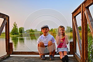 Cute children, little boy and girl are sitting on pier in the summer evening. Two siblings. Golden hour by the lakeside. Happy chi