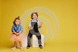 Cute children posing over a yellow background. Sad little girl and happy boy in casual outfits on the yellow background