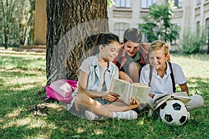 Cute classmates reading book and smiling while sitting on lawn under tree