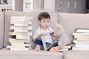 Cute clever funny little baby boy with thoughtful serious face expression holding glasses in hands reading book sitting on sofa wi