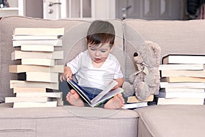 Cute clever little baby boy keen about reading book sitting on sofa with teddy bear toy and piles of books