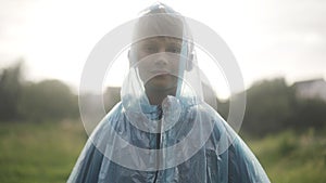 Cute confident boy zipping raincoat and looking at camera standing on rainy summer day outdoors. Portrait of cute