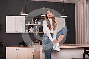 Cute cool young woman with long red hair in trendy youth casual blue-white wear posing sitting on a wooden table in the cook room