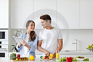 Cute couple cooking in kitchen, man chopping vegetables while his girlfriend is using a digital tablet looking for a recipe.