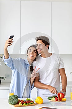 Cute couple cooking in kitchen, man chopping vegetables at this time his girlfriend taking a selfie on a smartphone together.