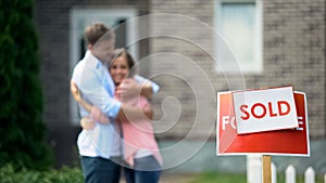 Cute couple hugging and smiling, celebrating purchase of great house, signboard