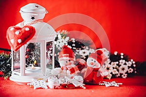 Cute couple of little snowmen is standing near the white fairy lantern with a toy heart on it and decorated fir tree branch.