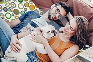 Cute couple lying in a hammock with a dog, funny white bulldog relaxing with a hipster young family on a beach, family portrait