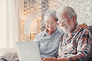 Cute couple of old people sitting on the sofa using laptop together shopping and surfing the net. Two mature people wearing