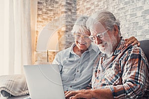 Cute couple of old people sitting on the sofa using laptop together shopping and surfing the net. Two mature people wearing