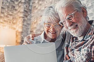 Cute couple of old people sitting on the sofa using laptop together shopping and surfing the net. Two mature people wearing