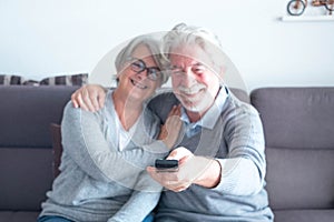 Cute couple of two caucasia mature seniors o the sofa watching  a film or something in the TV - man holding the remote control