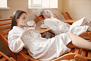 Cute couple in white bathrobes laying on a sunbeds indoors and drink a tea