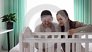 A cute couple of young parents are hugging near the baby's cradle and looking tenderly at the baby. Happy family and