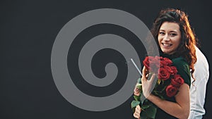 Cute curly brunette girl standing, smelling red roses. Courier is walking on the background with clipboard.