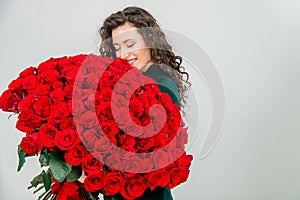 Cute curly brunette girl standing, smelling red roses over white background.
