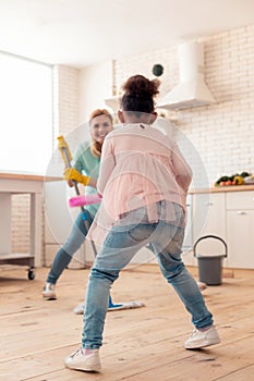 Cute curly daughter mopping floor with mother and dancing
