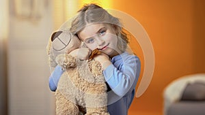Cute curly-haired blond girl hugging teddy bear and smiling at camera, happiness