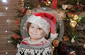 Cute curly-haired boy in a red Santa hat at the Christmas tree