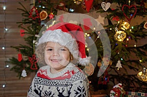 Cute curly-haired boy in a red Santa hat at the Christmas tree