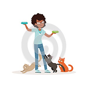 Cute curly-haired girl feeding homeless cats. Volunteer holding bowls with milk. Social help. Teen in glasses, blue t