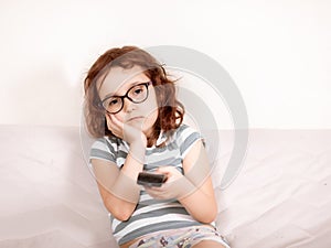 Cute curly little girl kid child in a eyeglasses bored watching tv at home with remote control in a hands