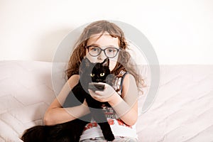 Cute curly little girl kid child in a eyeglasses watching TV with kitty cat best friend at home with remote control in a hands