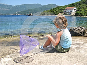 Cute curly toddler girl with butterfly net for catching fish sitting on the seashore, lookig at sea