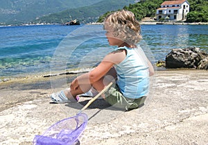 Cute curly toddler girl with butterfly net for catching fish sitting on the seashore, lookig at sea