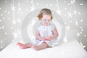 Cute curly toddler girl in a white dress on bed between Christmas lights