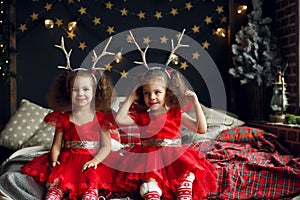 Cute curly twins girl sitting on the bed in decorated xmas room, on Christmas evening 2018 near New Year fir tree with