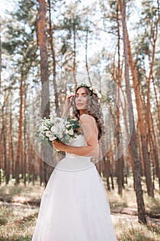 A cute curly woman in a white wedding dress with a wedding bouquet and wreath in her hair standing back to the camera in nature.