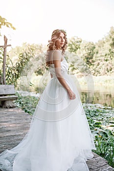 A cute curly woman in a white wedding dress with a wedding bouquet and wreath in her hair standing back to the camera in nature.